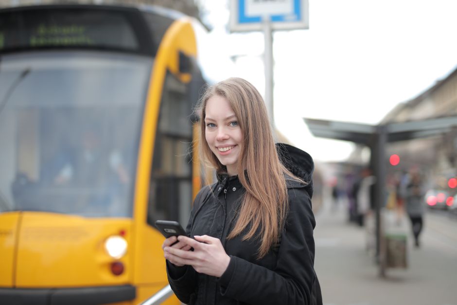 Mobilidade Urbana — Smiling woman texting on phone at tram station in city street setting.
