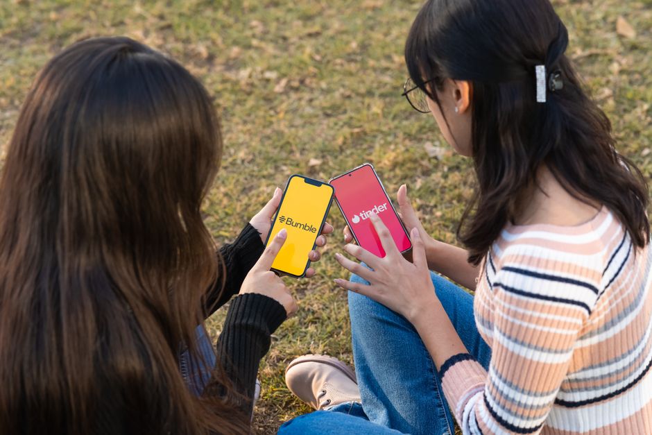 Mobilidade Urbana — Two women using dating apps Bumble and Tinder on smartphones in a park setting.